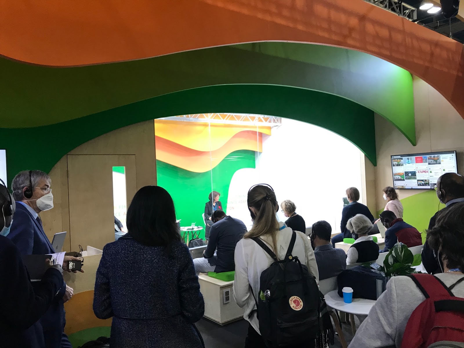 A small crowd of observers listens to headphones as they stand outside a glass-walled room in which panelists speak A small crowd of observers listens to headphones as they stand outside a glass-walled room in which panelists speak