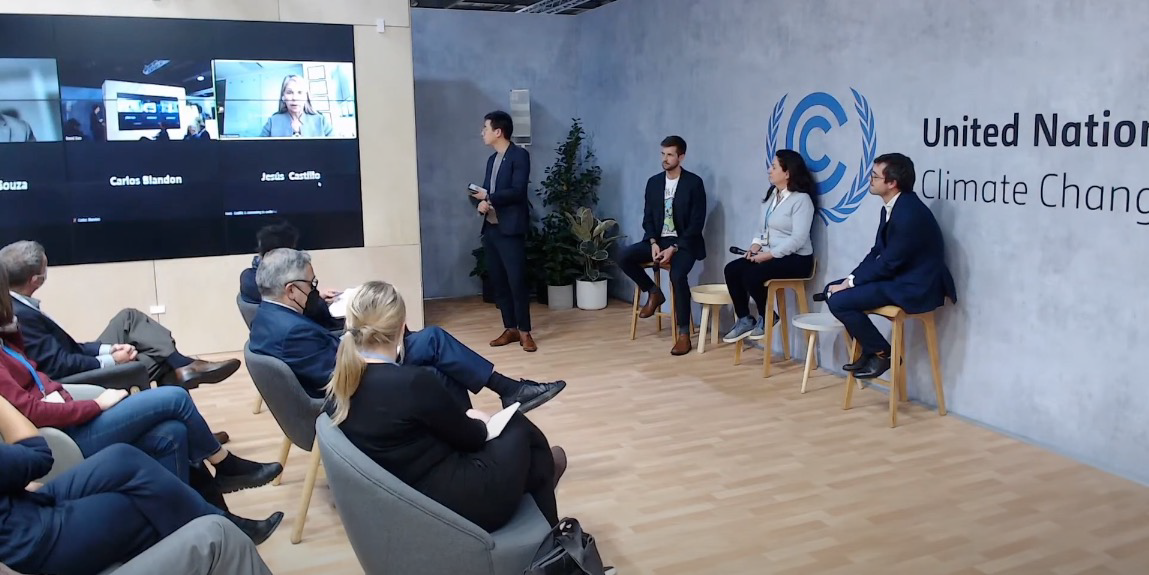 A moderator stands between three seated live panelists and a large screen showing several virtual panelists A moderator stands between three seated live panelists and a large screen showing several virtual panelists